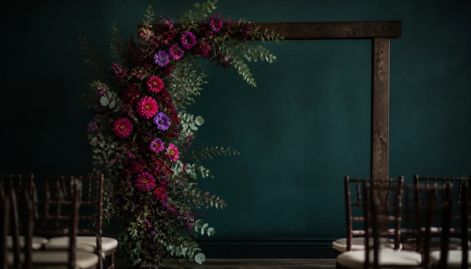 Asymmetric wedding ceremony arch of jewel-toned dahlias, garden roses and anemones climbing a dark wooden frame